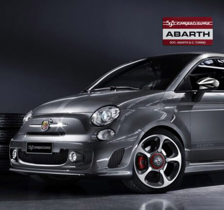 Abarth 595 Competizione grigio campovolo con a fianco degli pneumatici e sullo sfondo un muro a scacchi rosso e bianco